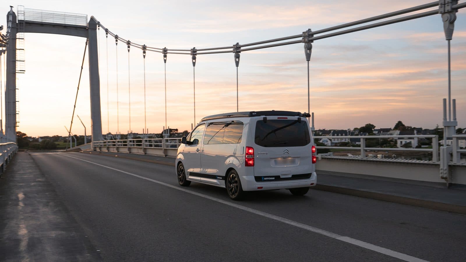 Van aménagé roulant sur un pont suspendu au coucher du soleil